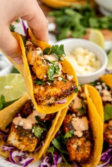 Someone is lifting a fish taco from a platter filled with the same tacos.