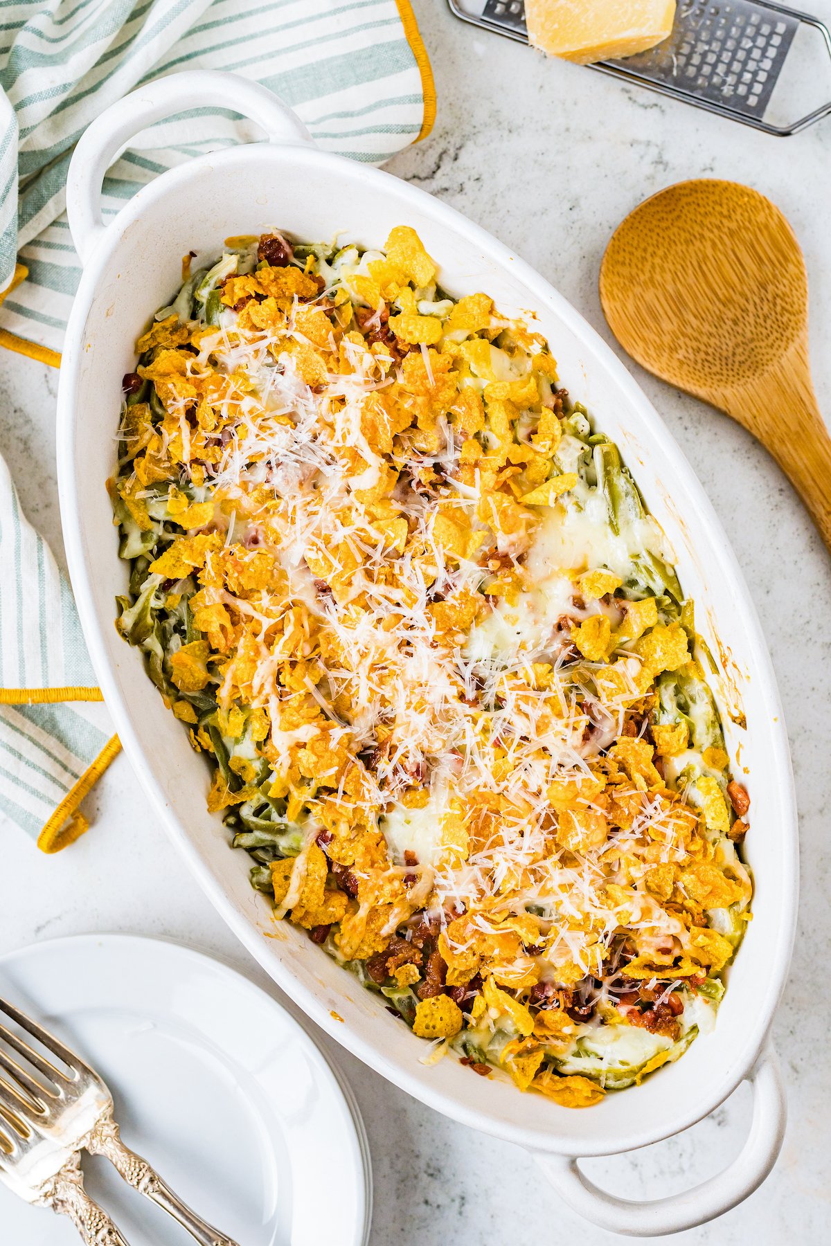 Cheesy Creamed Green Beans are a satisfyingly cheesy green bean casserole made with sour cream and topped with cheese, bacon, and cornflakes. An oval casserole dish with a green bean casserole in it, topped with cornflakes and Parmesan.