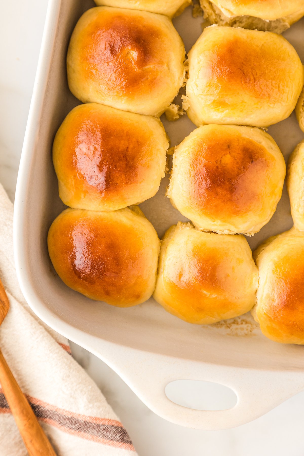 Golden brown Hawaiian rolls are arranged closely together in a white baking dish. A striped kitchen towel and a wooden spoon are partially visible next to the dish on a light surface.